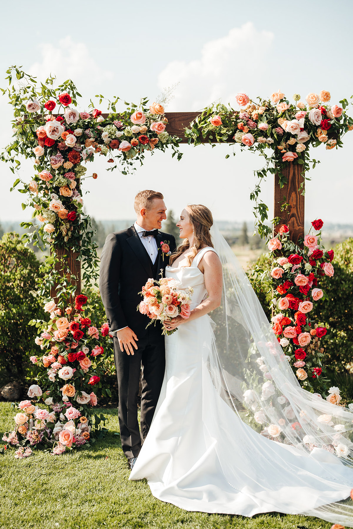 A bride a groom stand in front of an arbor covered in flowers
